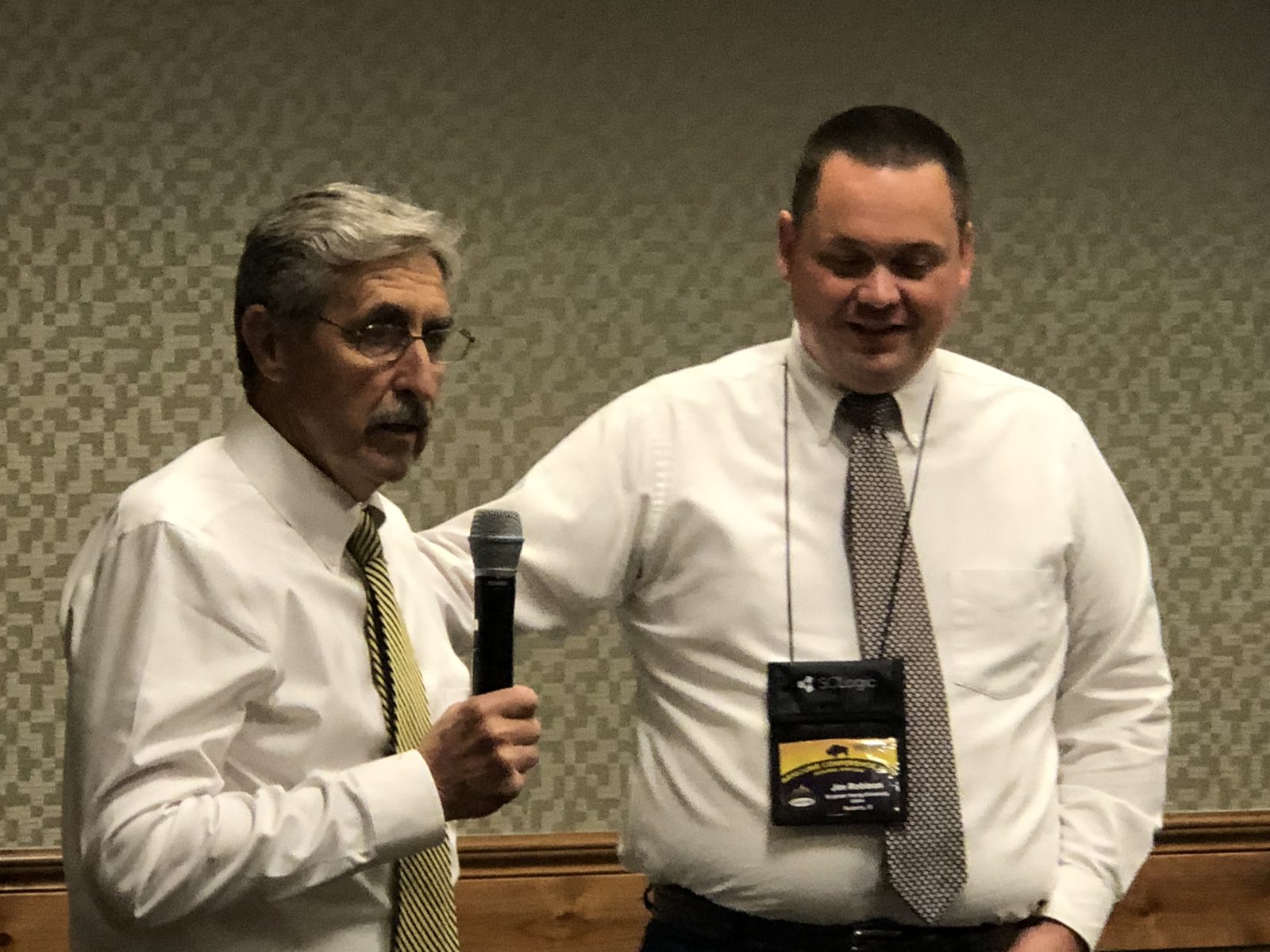 Two men in white shirts and ties, one holding a microphone.