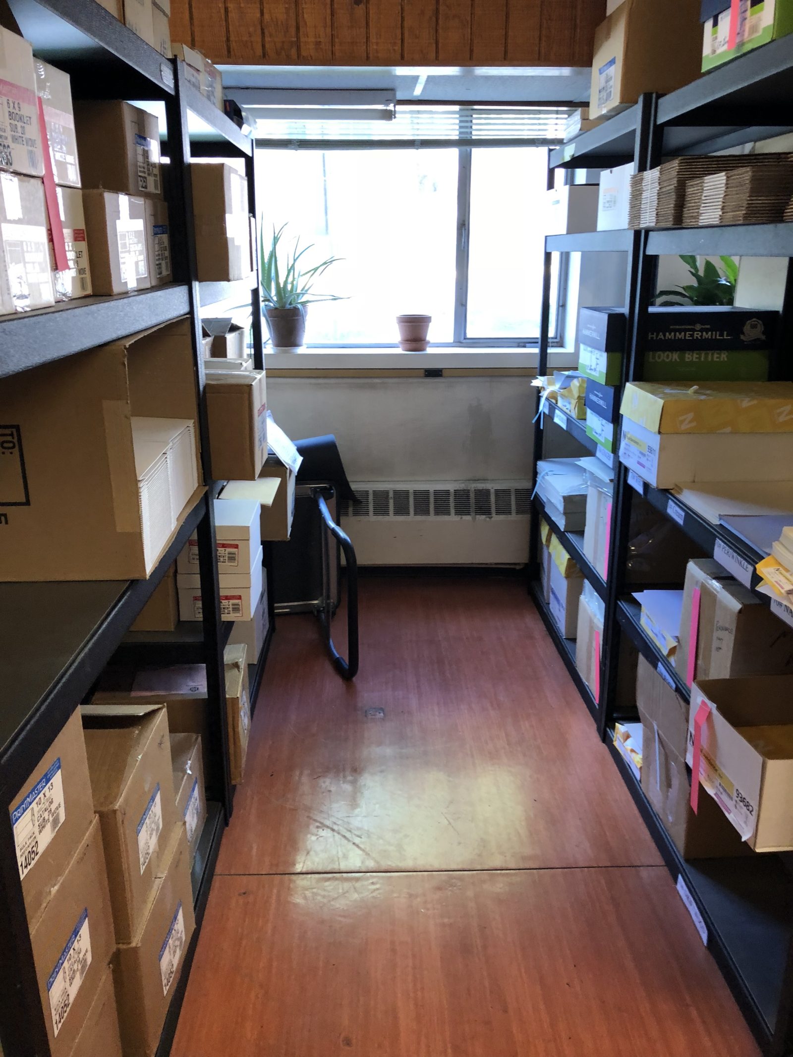 A narrow storage room with shelves full of boxes and a desk by the window.