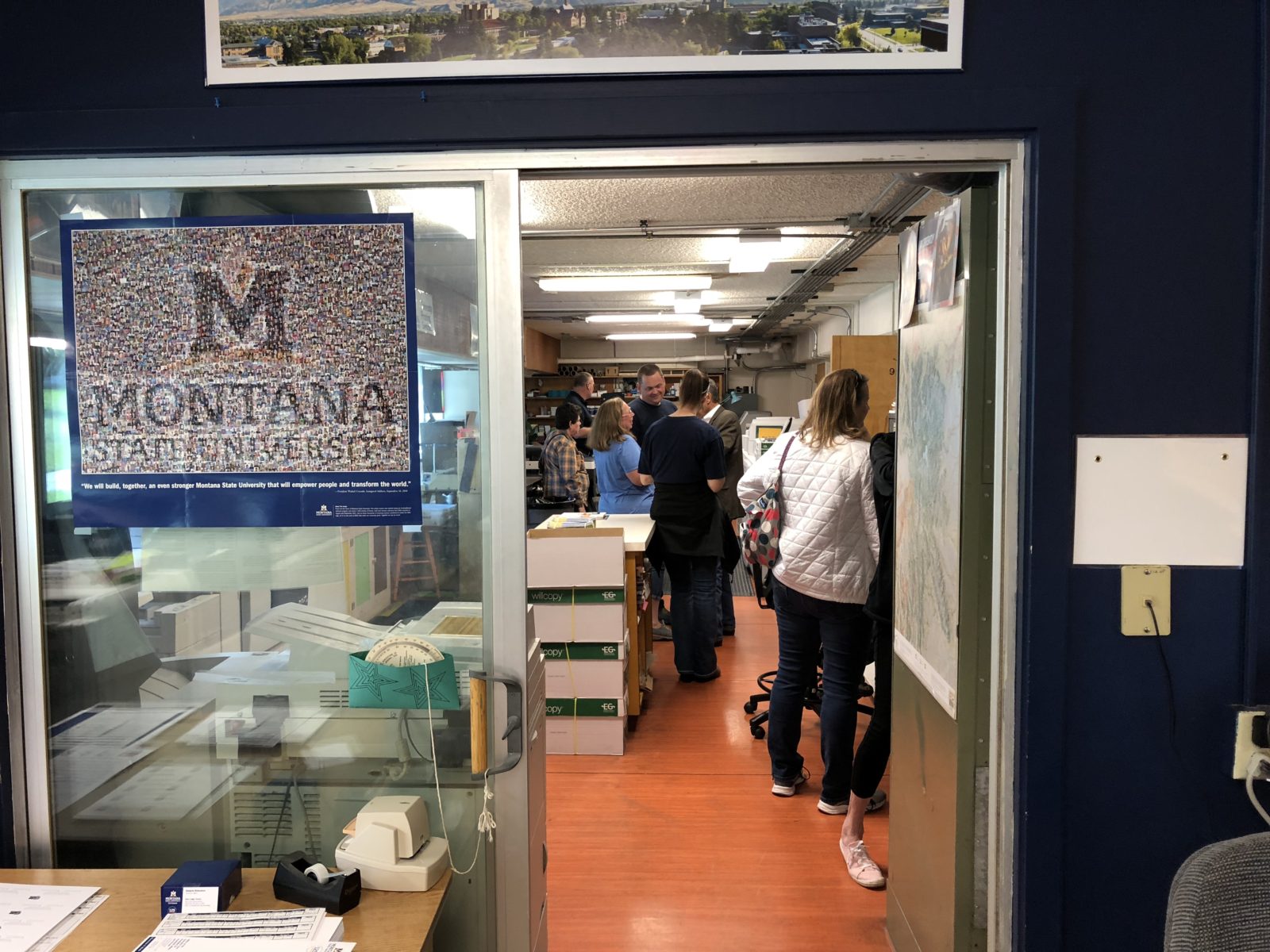 People wait in line inside a narrow hallway with a banner on the door.
