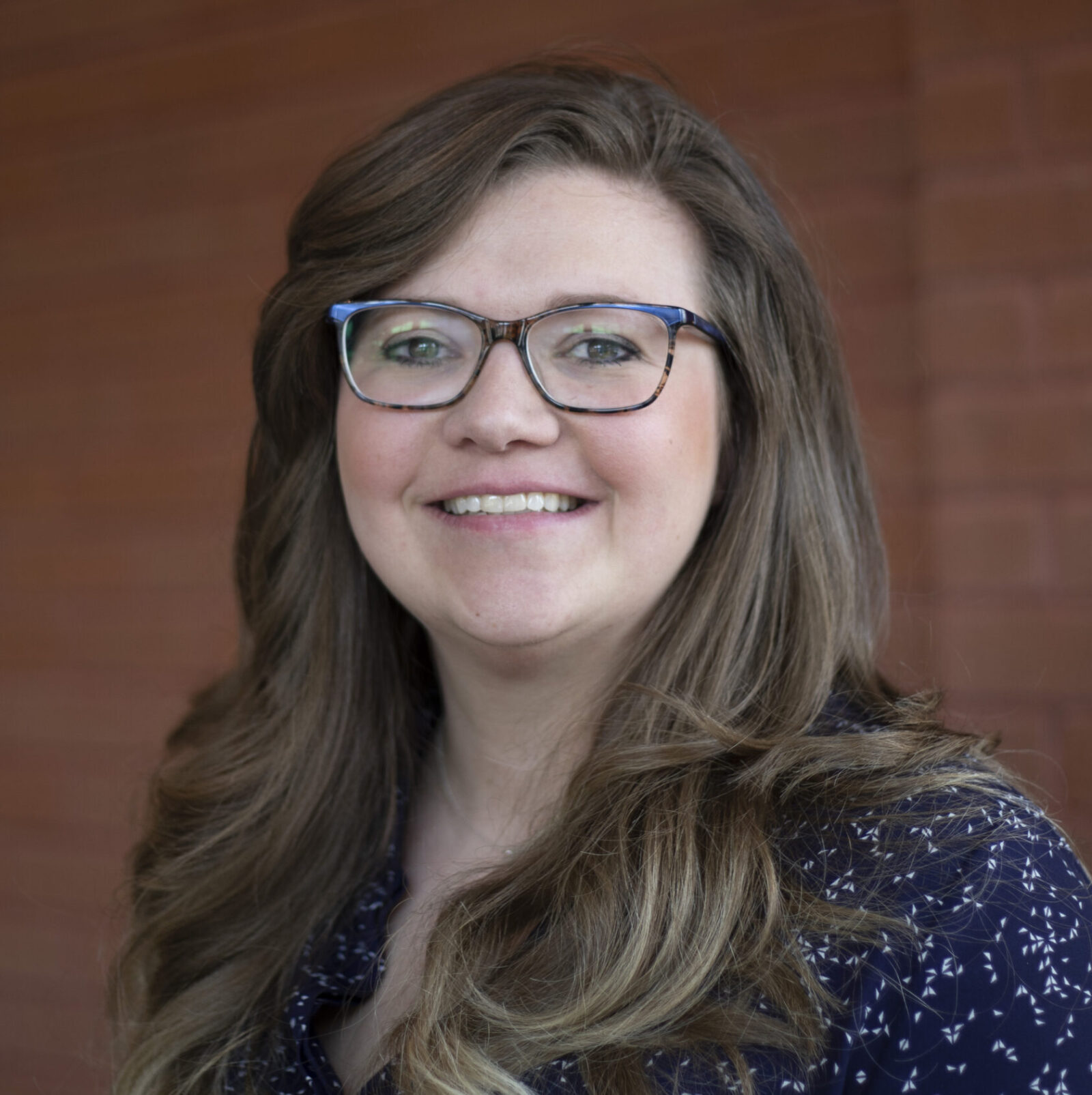 Smiling woman with glasses and wavy hair in front of a brick wall.